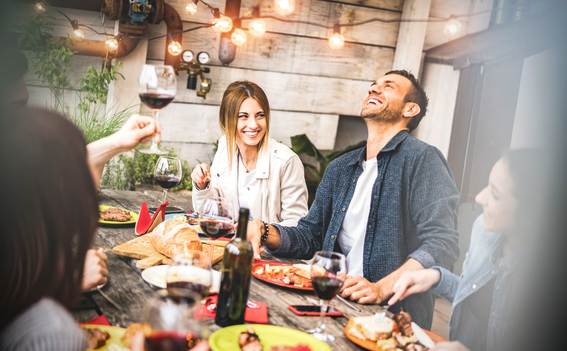 Group of friends laughing at a dinner table with food and wine on an outdoor patio with string lights and plants.